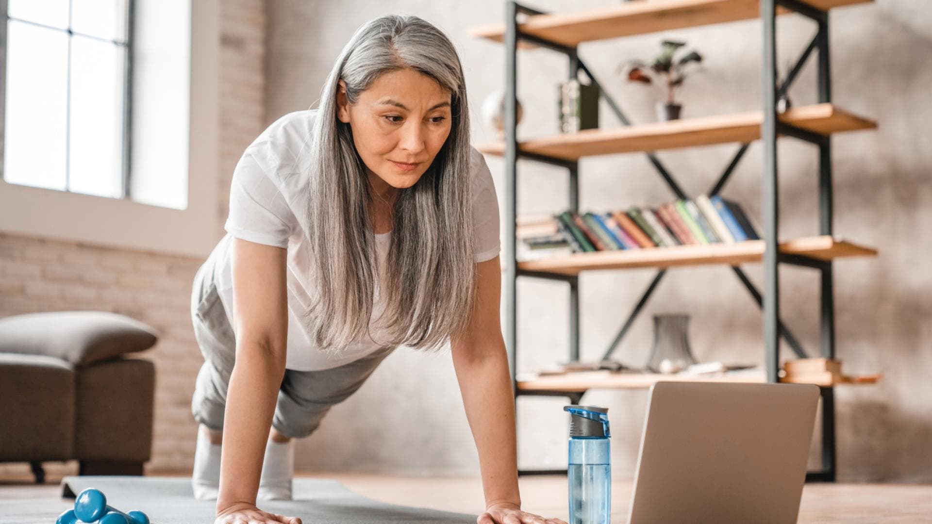 Regelmäßige körperliche Aktivität fördert den Knochenaufbau und wirkt Osteoporose entgegen. Frau im mittleren Alter macht Liegestütz zu Hause vor Laptop