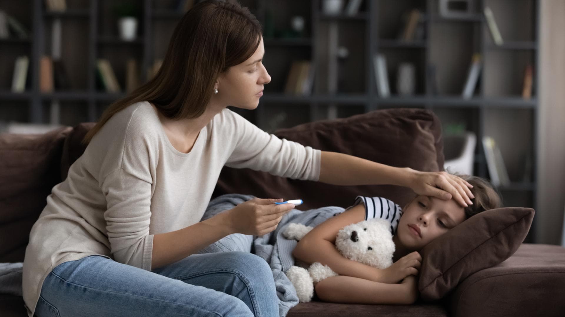 Kind liegt krank auf dem Sofa, während die Mama Fieber misst