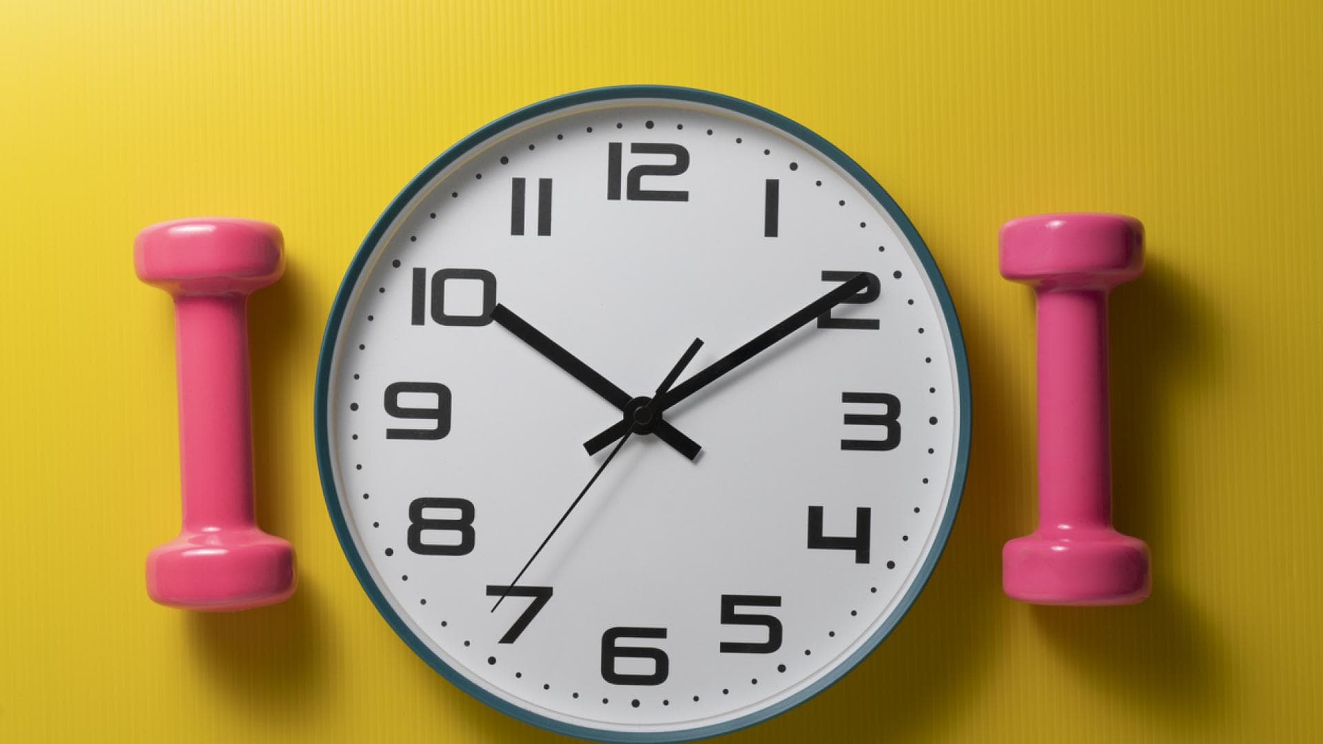 Analoge Wanduhr vor gelbem Huntergrund mit pinken Hanteln an der Seite 