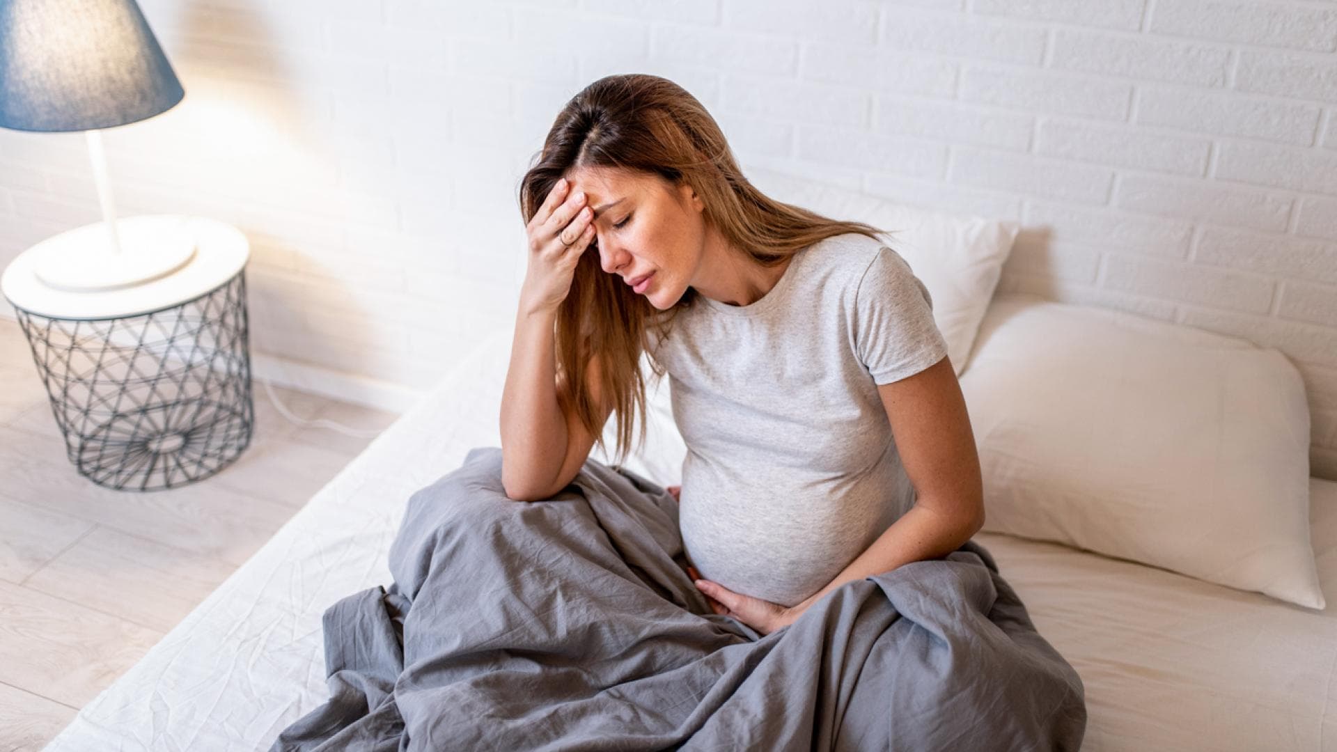 Schwangere sitzt im Bett und hält sich Kopf und Bauch