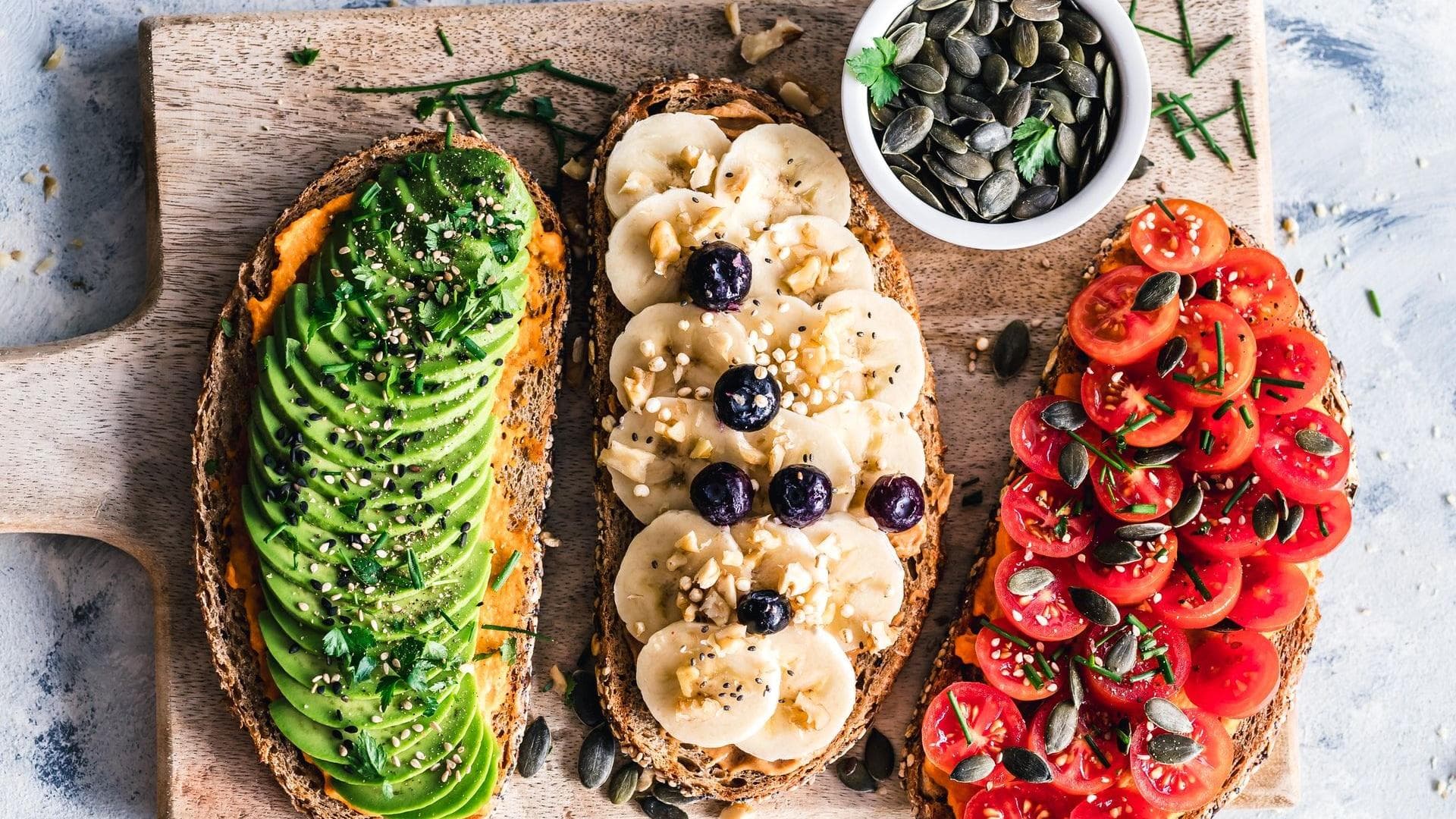 Vegane Ernährung ist abwechslungsreich Drei belegte Stullen nebeneinander auf einem Holzbrett mit Avocado, Banane und Tomate