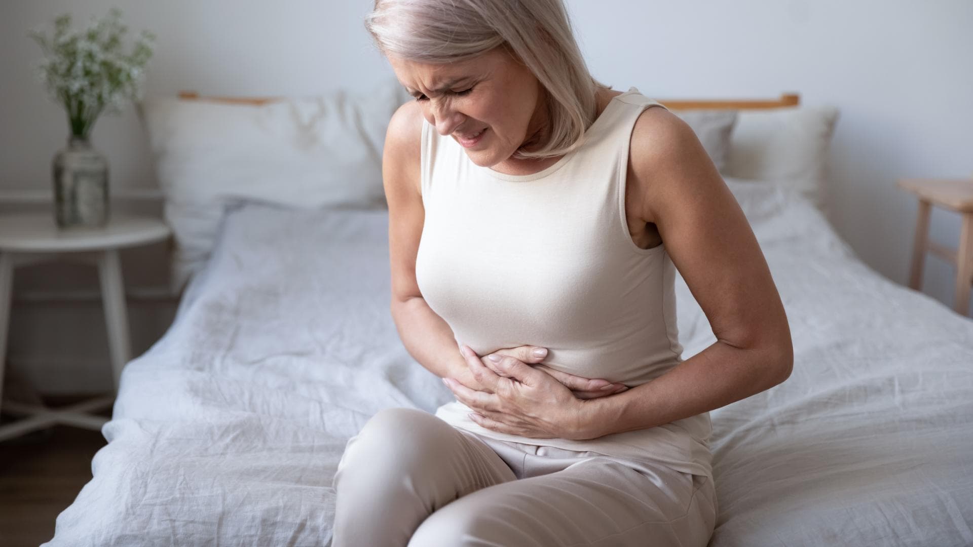 Reizdarm wird zur Volkskrankheit. Kann man vorbeugen? Frau sitzt auf Bett und hält sich mit schmerzverzerrtem Geischt den Bauch