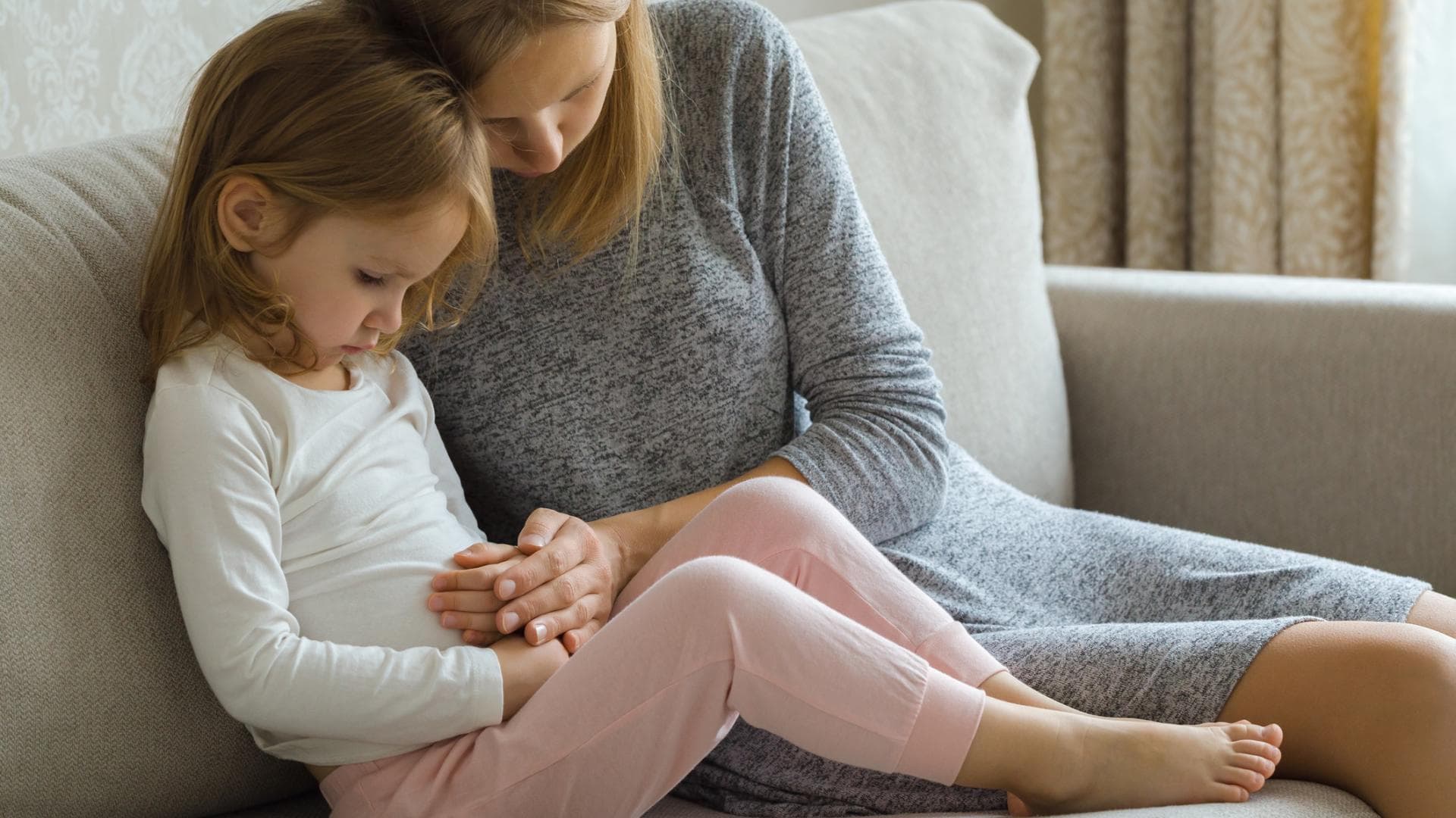 Mädchen sitzt mit Bauchschmerzen auf der Couch und wird von der Mutter getröstet. Bauchschmerzen bei Kindern: Mädchen sitzt mit Bauchschmerzen auf der Couch und wird von der Mutter getröstet.