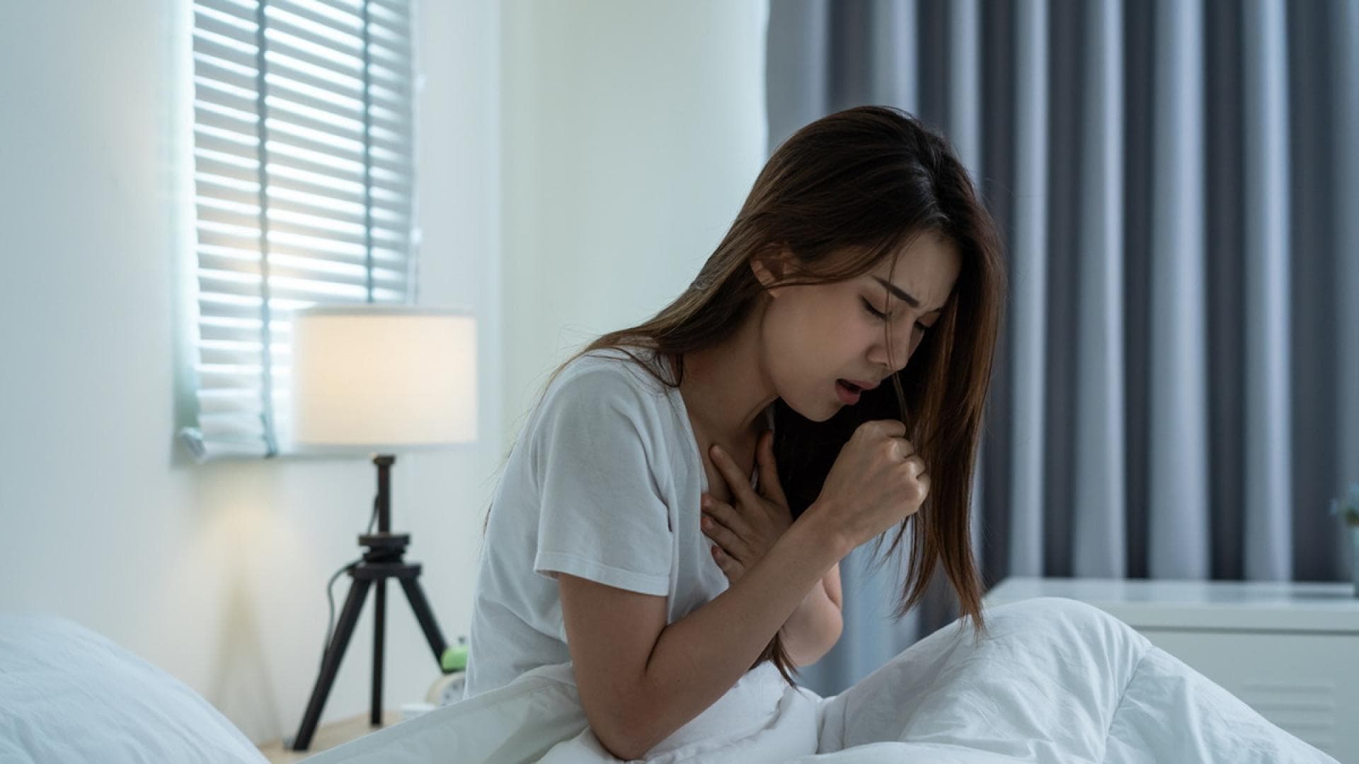 Reizhusten, sobald Sie sich hinlegen, kann den letzten Nerv rauben Junge Frau sitzt im Bett auf und hustet in ihre Faust