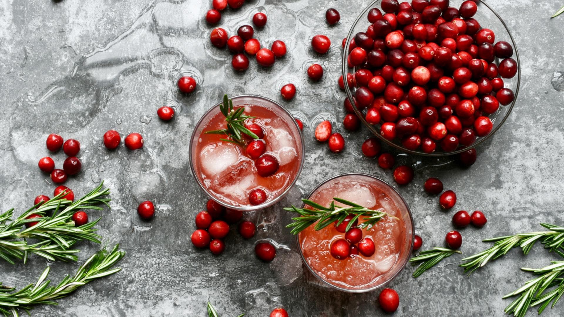 Cranberrysaft hat viele gesundheitliche Vorteile Draufsicht auf eine Schale mit frischen Cranberrys und zwei Gläsern Cranberrysaft mit Eiswürfeln und Rosmarin