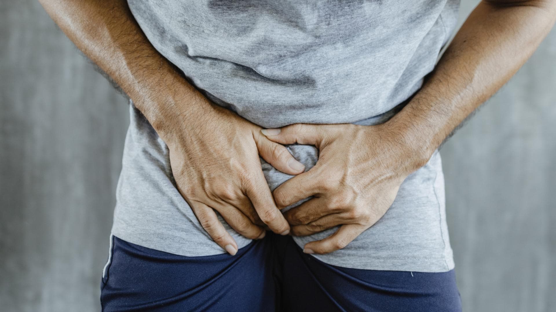 Eine Vorhautentzündung kann schmerzhaft sein Nahaufnahme eines Mannes in kurzer blauer Hose mit grauem Shirt, der sich vor Schmerzen in den Schritt greift