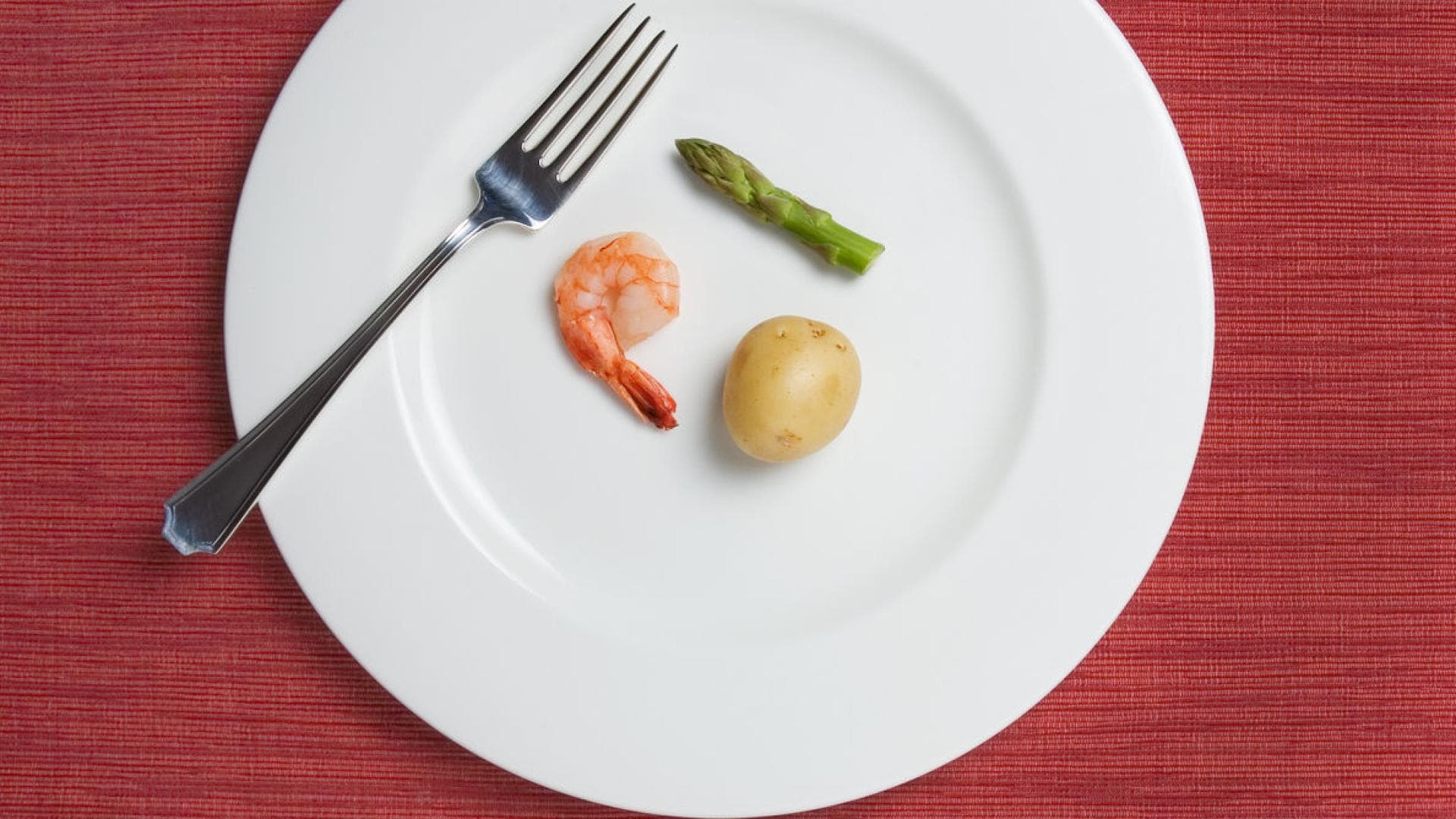 Großer Teller mit einer kleinen Kartoffel, einem Shrimp und einem Stück grünem Spargel darauf Großer Teller mit einer kleinen Kartoffel, einem Shrimp und einem Stück grünem Spargel darauf