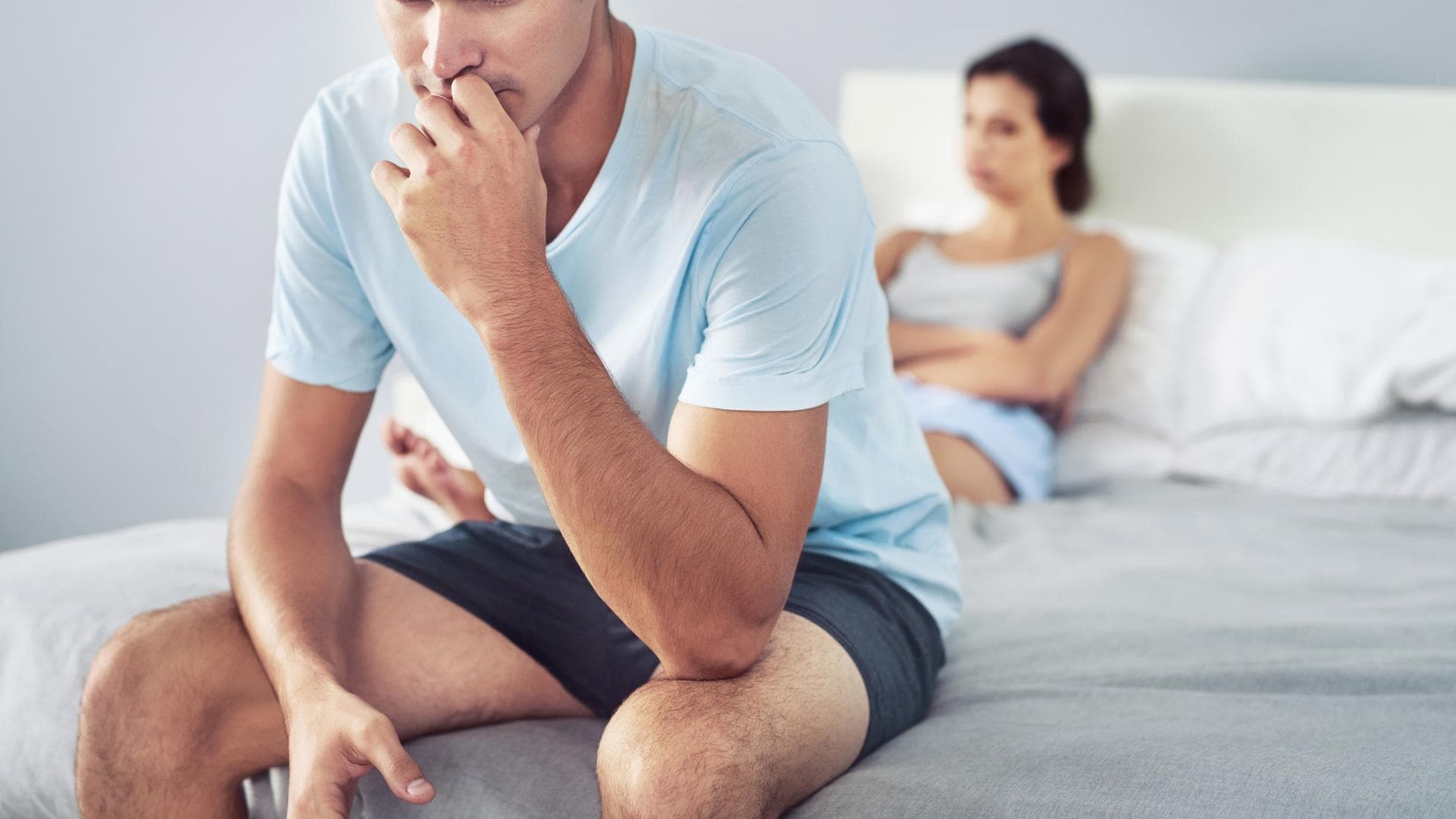 Mann sitzt auf dem Bett und ist verzweifelt mit Frau im Hintergrund Mann sitzt auf dem Bett und ist verzweifelt mit Frau im Hintergrund