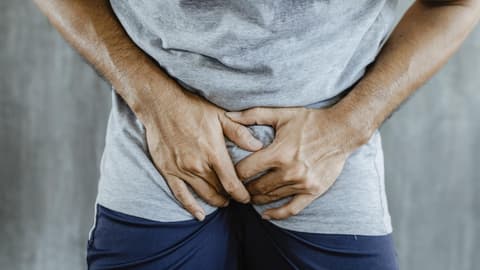 Nahaufnahme eines Mannes in kurzer blauer Hose mit grauem Shirt, der sich vor Schmerzen in den Schritt greift