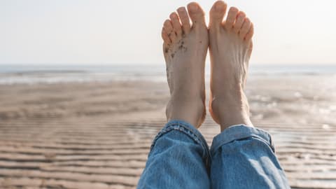 Eine Frau hält ihre nackten Füße hoch. Sie liegt am Strand. Ihre Fußnägel sind naturbelassen.