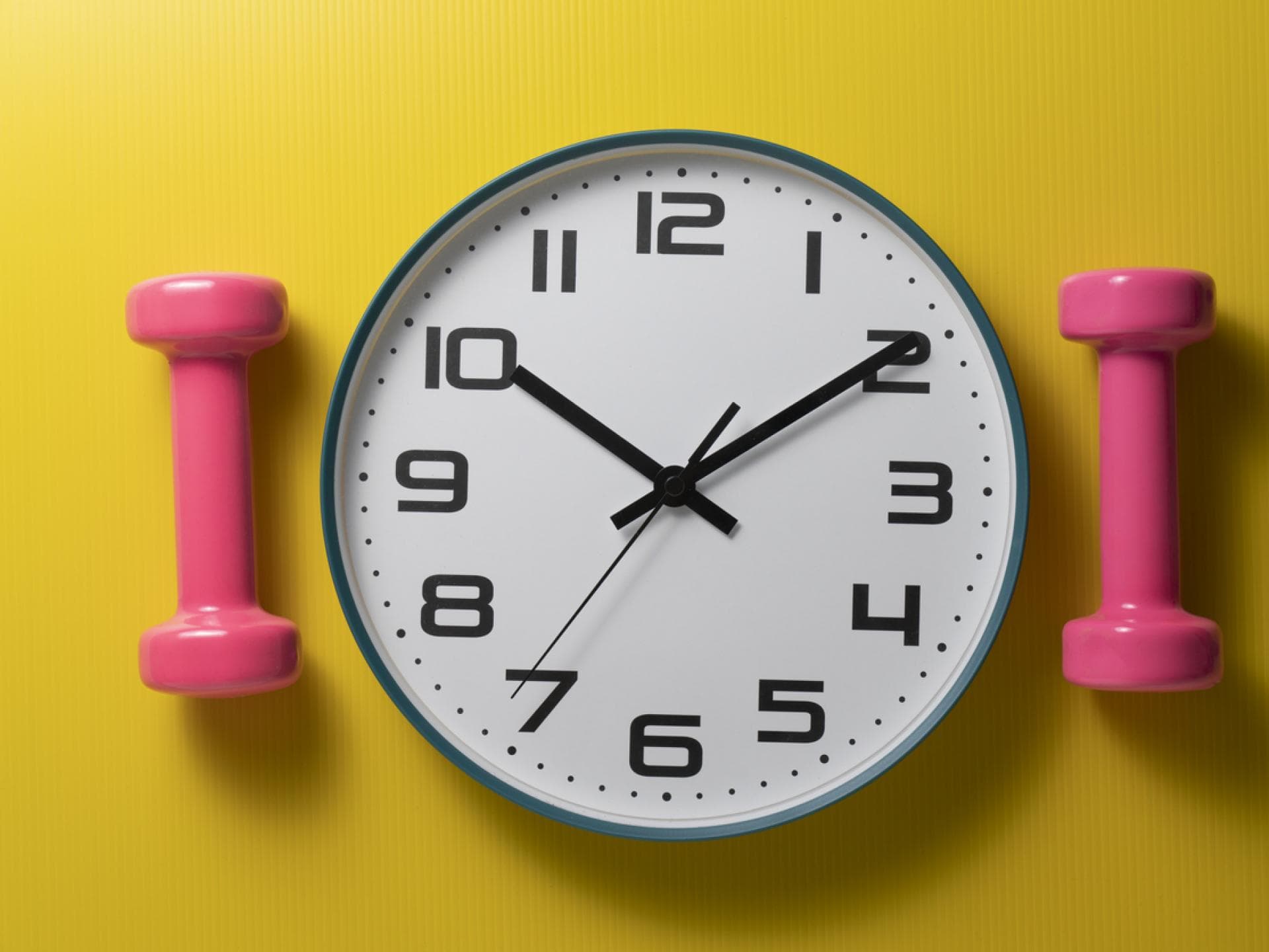 Analoge Wanduhr vor gelbem Huntergrund mit pinken Hanteln an der Seite 