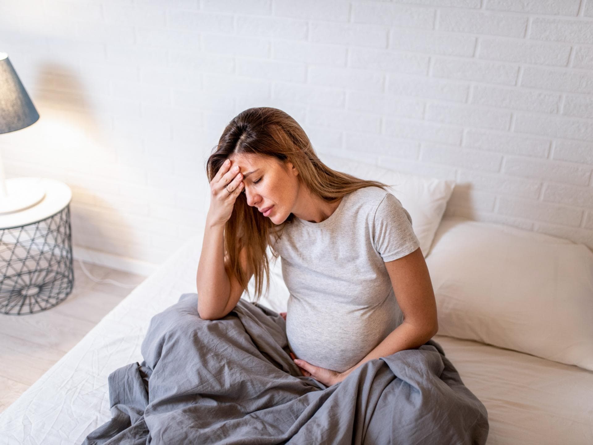 Schwangere sitzt im Bett und hält sich Kopf und Bauch