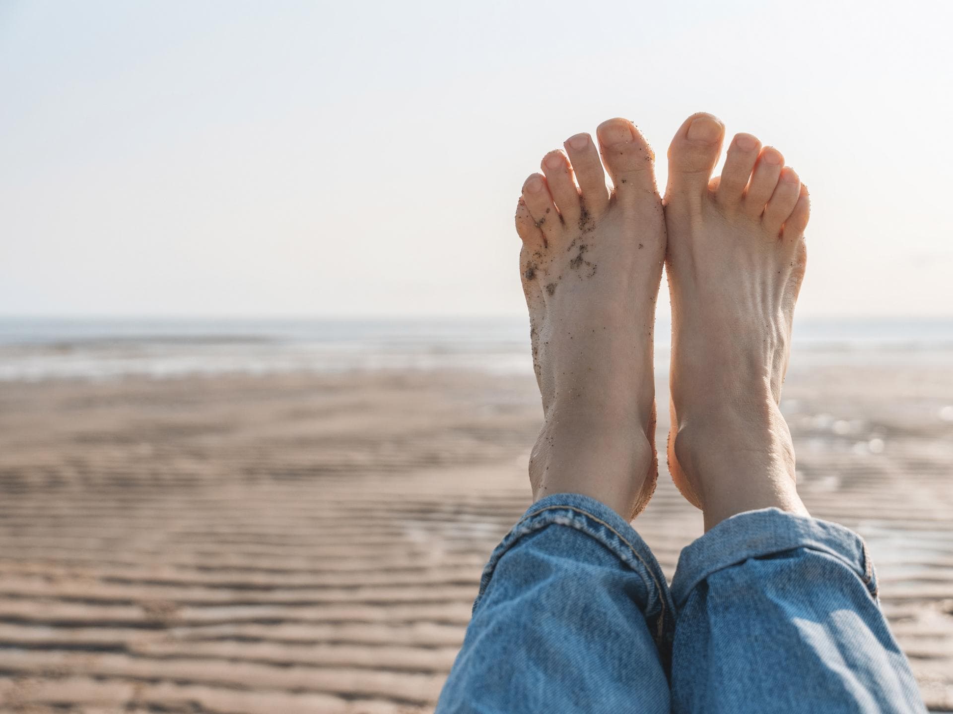 Eine Frau hält ihre nackten Füße hoch. Sie liegt am Strand. Ihre Fußnägel sind naturbelassen.