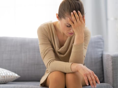 Frau sitzt gebeugt auf einem Sofa und hält sich die Hand vor den Kopf