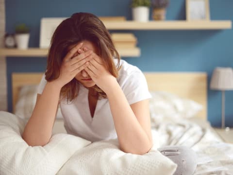 Frau mit Erschöpfung sitzt im Bett mit Händen vor dem Gesicht