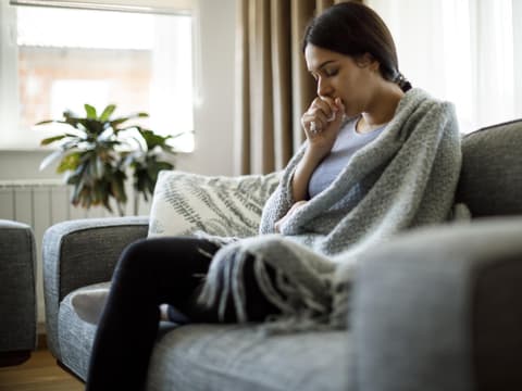 Frau mit Erkältung sitzt auf Couch und hustet in ihre Hand