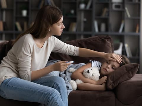 Kind liegt krank auf dem Sofa, während die Mama Fieber misst