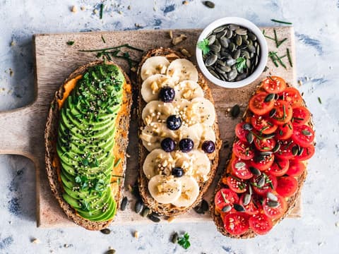Drei belegte Stullen nebeneinander auf einem Holzbrett mit Avocado, Banane und Tomate