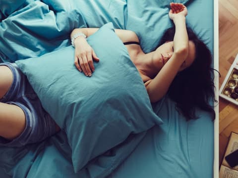 Frau mit Kopfschmerzen liegt im Bett, hat Kissen auf dem Bauch und den Arm über das Gesicht gelegt