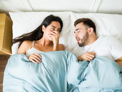 Frau liegt neben ihrem Freund im Bett und hält sich die Hand vor den Mund