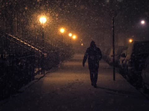Person geht bei Schneefall am Abend durch dunkle von Laternen beleuchtete Straße