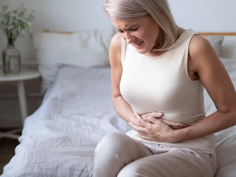 Frau sitzt auf Bett und hält sich mit schmerzverzerrtem Geischt den Bauch