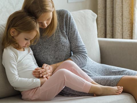 Bauchschmerzen bei Kindern: Mädchen sitzt mit Bauchschmerzen auf der Couch und wird von der Mutter getröstet.