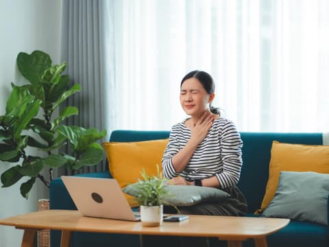 Frau sitzt im Wohnzimmer am Laptop und fasst sich an den schmerzenden Hals