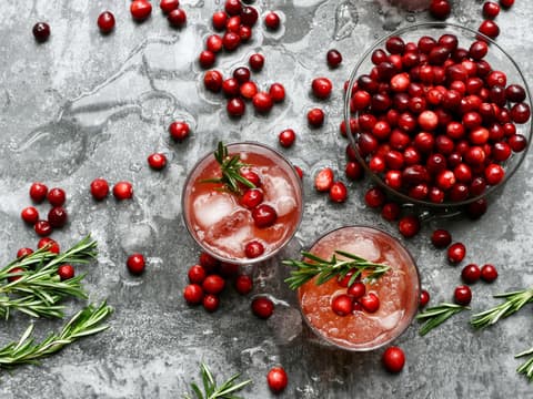 Draufsicht auf eine Schale mit frischen Cranberrys und zwei Gläsern Cranberrysaft mit Eiswürfeln und Rosmarin