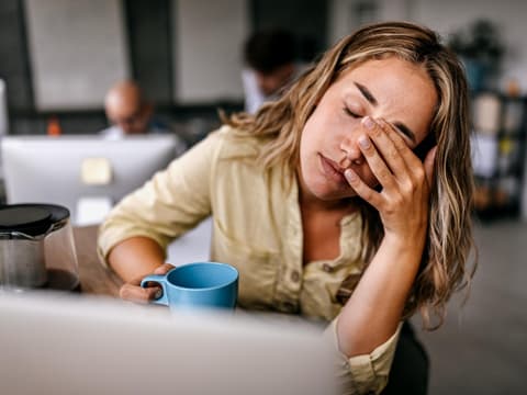 Müde Frau sitzt mit geschlossenen Augen und Kaffeebecher vor dem Laptop