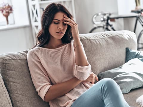 Frau hat einen Schwächeanfall und sitzt auf Sofa mit Hand an der Stirn