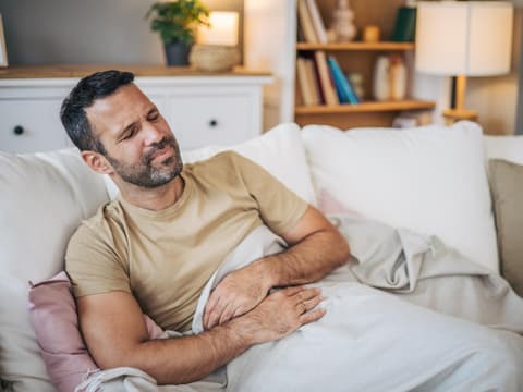 Mann mit Magen-Darm-Grippe hält sich schmerzenden Bauch auf dem Sofa