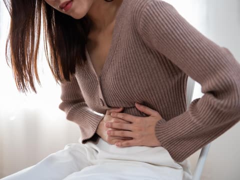Frau in braunem Strickpulli und weißer Hose mit Darmbeschwerden hält sich den Bauch