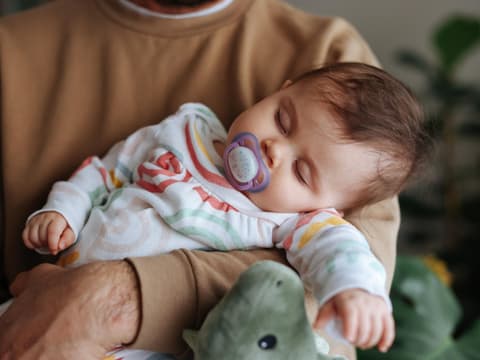 Ein Baby schläft mit einem Schnuller auf dem Arm des Vaters.