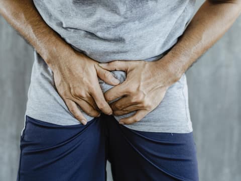 Nahaufnahme eines Mannes in kurzer blauer Hose mit grauem Shirt, der sich vor Schmerzen in den Schritt greift