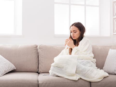 Frau sitzt frierend in einer Decke auf dem Sofa
