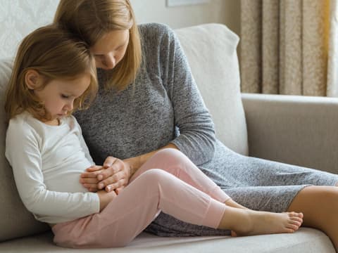 Kind sitzt mit Mutter auf dem Sofa und hält sich den Bauch