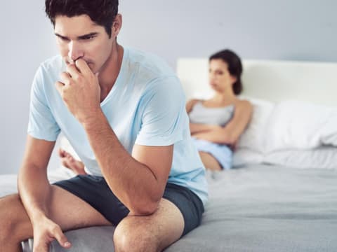 Mann sitzt auf dem Bett und ist verzweifelt mit Frau im Hintergrund