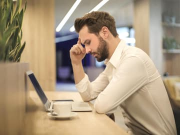 Mann sitzt vor Laptop und Kaffeetasse und fasst sich an die Schläfe Mann sitzt vor Laptop und Kaffeetasse und fasst sich an die Schläfe
