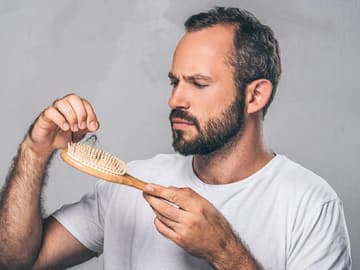 Mann Bürste Tipps gegen Haarausfall Mann zieht Haare aus Haarbürste und sucht nachdenklich nach Tipps gegen Haarausfall